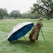 A golf umbrella in navy blue and white is open on a grassy golf course next to a brown golf bag filled with clubs.