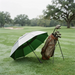 A golf umbrella in green and white lies on its side on a grassy golf course, next to a brown golf bag filled with clubs.