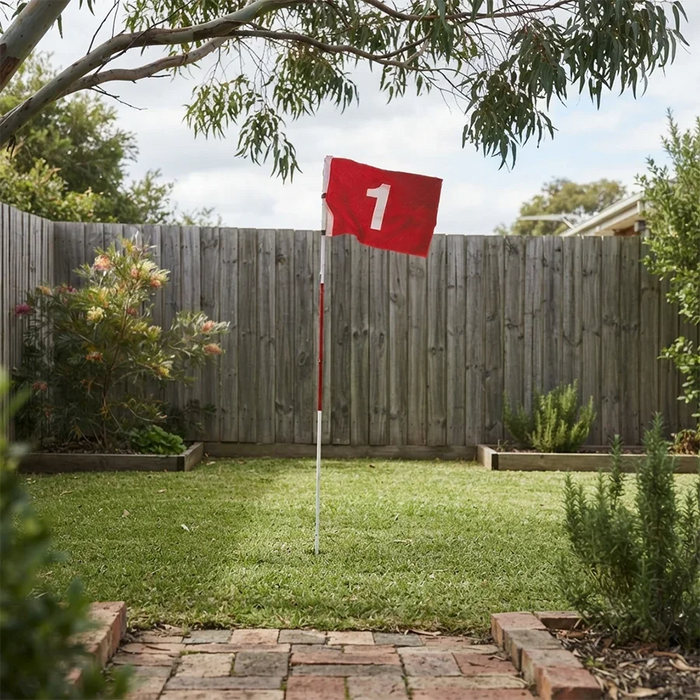 A red flag with the number 1 on a white pole stands on a grassy backyard lawn in front of a wooden fence, with garden plants, trees, and a brick path visible.