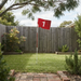 A red flag with the number 1 on a white pole stands on a grassy backyard lawn in front of a wooden fence, with garden plants, trees, and a brick path visible.