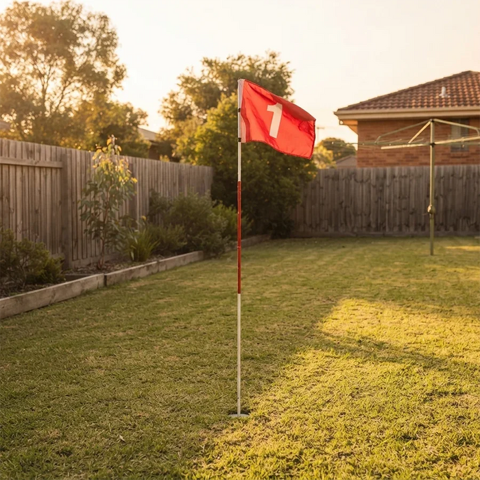 A red golf flag with a white number 1 stands in a grassy backyard at sunset, with a wooden fence, trees, and part of a house visible in the background.