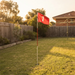 A red golf flag with a white number 1 stands in a grassy backyard at sunset, with a wooden fence, trees, and part of a house visible in the background.